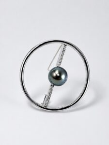 Silver circular hoop jewelry with a diagonal diamond-studded bar holding a dark grey-blue pearl centerpiece.